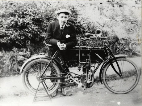 Photograph of a young man wearing a suit and a cap and smoking a pipe, leaning on a bicycle, which is on a stand; behind him are bushes; on the front mudguard of the bicycle are the letters J-910 and on the cross bar the initials N. S. U.; he has been identified as Dick Walker, 1907