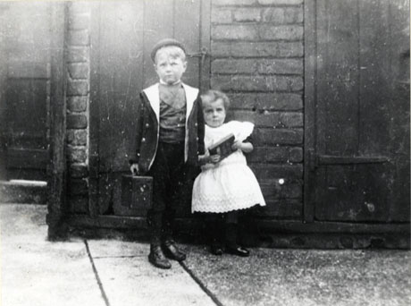 Photograph of a small boy, aged approximately five years, wearing a jacket and cap, standing with a small girl aged, approximately three years, wearing a broderie anglaise pinafore and carrying a slate; they are posed in front of what appears to be the door of an outside lavatory; they have been identified as Winnie and Joe Richardson