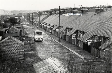 Photograph of the back lanes of St. Andrews Terrace, Cold Hesledon, showing an unmetalled road running away from the camera and the back yards of a row of terraced houses on the right of the picture; a further road in the right beyond St. Andrews Terrace leads towards open country; on the left hand side of the picture the sides of sheds can be seen on the opposite side of the road from the houses in St. Andrews Terrace; two caravans and two cars can be seen parked in the distance and an ice cream van is moving along the road