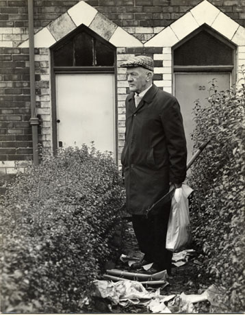 Photograph of a middle-aged man outside the door of a house of brick with bands of light-coloured brick across the facade and round the doorway; he is wearing a raincoat, tie and cap; he is holding a spade and plastic carrier bag and paper and plastic rubbish is lying at his feet; he is standing between two low but overgrown hedges; he has been identified as Mr. Avery outside Hesledon Terrace