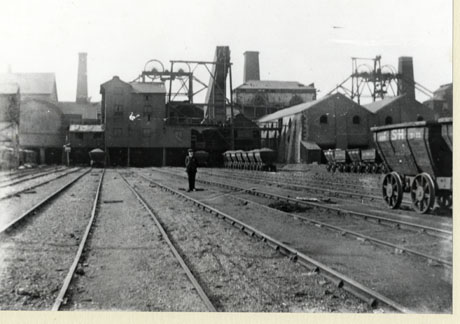 Photograph showing rail tracks running away from the camera towards colliery buildings, behind which winding gear and chimneys can be seen ; a man is standing between the tracks in the middle distance; six trucks are on the tracks in the distance, four trucks in the middle distance and one truck in the foreground to the right; the photograph has been identified as depicting Murton Colliery
