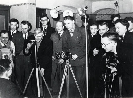 Photograph of fourteen men standing in a room with tripods, cameras, and lights looking at a man at the front of the photograph, who can be seen only from the rear; the men have been identified as members of the Murton Camera Club