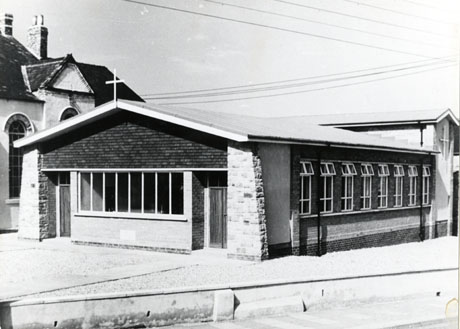 Photograph of the exterior of a low building attached to a church, most likely a church hall; the building is in the style of the 1960s-1970s; the front and side of the building can be seen; behind the low building a higher building with a cross on its side can be seen; also behind the building an older building with a gable and long round-headed widows can be seen; the photograph has been identified as St. Paul's Church, Murton
