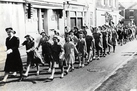 Photograph of a procession of eight young girls in ordinary clothes and approximately twenty girls in uniform, followed by boys, who appear to be in the uniform of the boy scouts and the cubs; the procession is lead by a woman in uniform and is going past the facade of shops, one of which is Jeffery's; the procession has been described as St. John's Cadets; at the end of the buildings is The Colliery Inn