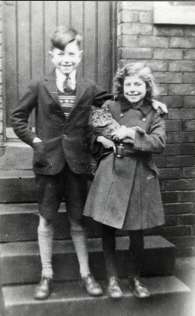 Photograph of a boy and a girl standing on steps leading from the door of a house; the boy, on the left of the two, is aged approximately ten years and is wearing a suit, pullover, tie, long socks, and brogues; the girl is wearing a double-breasted coat, dark stockings, and brogues; she is also carrying a Tabby cat; they have been identified as Irene and Lambton Laurie