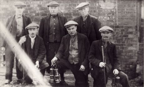 Photograph of six men wearing suits, waistcoats, scarves, and flat caps, standing in front of a brick wall, on which an indecipherable poster can be seen; four of the men can be seen to be carrying miners' lamps and two are carrying sticks; they have been identified as miners in Murton