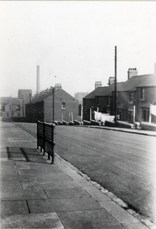 Photograph of a road running away from the camera downhill; on the left of the photograph is part of a pavement; across the road can be seen a block of houses with small gardens in which shopping lines can be seen; below that block is another, the side of which only can be seen; in the distance, buildings, possibly part of the colliery, can be seen along with a tall chimney; the photograph has been described as Murton's First Council Houses (Watt Street)