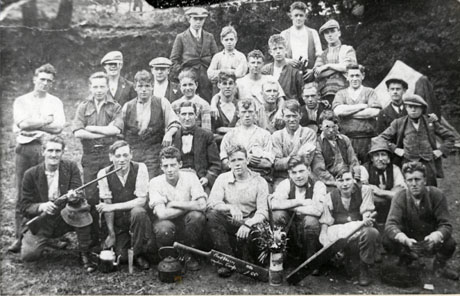 Photograph of a group of thirty-one men wearing working clothes posed in the open air; men on the front row are holding a rifle, a tea pot, a knife, a kettle; a man on the second row is holding an alarm clock; in front of the group are a vase of flowers and two cricket bats; on one of the bats is written : Hawthorn [ ] Group 1926