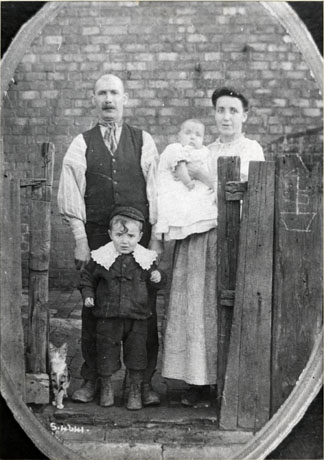 Photograph of a man wearing a waistcoat, shirt sleeves, and scarf, standing on the left of the picture; on the right of him is a woman in a crumpled gingham skirt and white blouse, carrying a baby in a long frock in her arms; in front of the man is a boy of approximately four years dressed in a suit with a large lace collar; both man and boy can be seen to be wearing boots; on the left of the man is a small tabby cat; the group are standing in the gateway of a rough fence; they have been identified as The Mullen Family