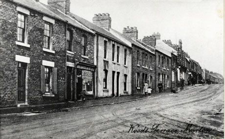 Postcard photograph entitled Woods Terrace, Murton, showing the surface of an unmade up road and, beyond it, terraced houses running uphill; one shop can be seen in the middle of the picture; further shops can be seen in the distance; indistinct figures can be seen on the pavement