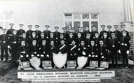 Photograph of thirty eight men in uniform, posed outside a building with mullioned windows; in front of the group are three shields and five canteens of cutlery; a printed caption to the photograph reads as follows: st. John Ambulance Brigade, Murton Colliery Division No. VI. District Murton, Co. Durham. 1912 Winners of Bronze Shield, Final Durham County Competition; Silver Shield and Medals, Final Durham, Northumberland and North-East Yorkshire Competition; Dewar Challenge Shield with Cases of Cutlery, Final Great Britain Competition; Heath Cup and Medals for Stretcher Work