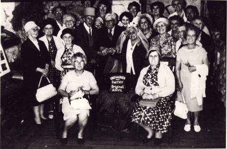 Photograph of a group of twenty two elderly people posed inside a building at Gretna Green; a man and a woman are pretending to be getting married: the man appears to be wearing a hat and the woman is wearing a wedding veil; in front of the pair is an object on which a sign can be seen reading as follows: Gretna Green Smithy Original Anvil; the other members of the party are grouped round the anvil; the photograph has been identified as Over Sixties Trip