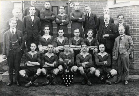 Photograph of twelve young men posed on the steps of a building, flanked by nine men in suits; ten of the young men are in football strip; in front of the group is a trophy cup and a shield with medals; they have been identified as Murton Footballers