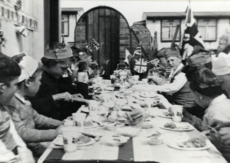 Photograph showing the surface of a table running away from the camera covered with a cloth, plates of food and mugs; children wearing party hats are sitting on either side of the table; at the end of the table the facades of houses can be seen; bunting and a Union Jack can be seen; the photograph has been identified as recording Coronation Celebrations 1953, Bevan Square, Murton