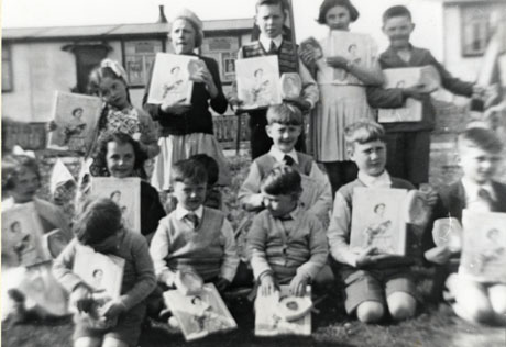 Photograph of fourteen children, aged between approximately four and eight years, kneeling and standing in two rows in the open air in front of very indistinctly seen houses; the children are each holding a photograph of Queen Elizabeth II; the picture has been described as Coronation Celebrations 1953 Bevan Square, Murton