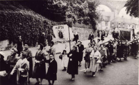 Photograph of a procession of women walking up North Road in the City of Durham; at the right of the picture the viaduct and a row of houses under the viaduct can be seen; the women are carrying a banner on which the words Murton Women's Section can be seen; the wall of Wharton Park can be seen behind the procession