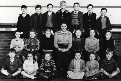 Photograph of nineteen children, aged approximately eight years, posed against the wall of a room with encaustic tiles on it; in the middle of the group a woman is seated; the photograph has been identified as Waterworks School Group