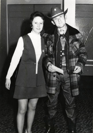 Photograph of a man dressed in a tartan jacket, shiny trousers and a stetson with a star on it, standing with a woman wearing a short skirt and long jerkin; they are photographed inside an unidentified building with patterned carpet; they have been identified as Jack Hedley and Girlfriend