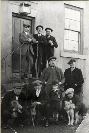 Photograph of five men crouching on the ground in front of a short flight of steps up the side of a building leading to a door; three of the men are holding dogs and the other two are standing with their hands in their pockets watching them; a small boy aged approximately three years is standing with the men; above the men on the ground, standing on the steps, are three men looking in the direction of the camera; all the men are wearing caps, and suits, typical of miners; two of the men have cigarettes in their mouths