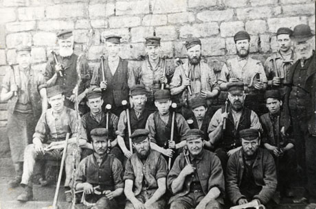 Photograph of a group of eighteen men posed in front of a stone wall; they are all wearing working clothes of waistcoat, shirt, scarf, and peaked cap, and are carrying hammers and pliers and other tools; three of the men are aged under twenty years; a man in a suit and bowler hat is standing at the right of the picture; they have been identified as Murton Colliery Blacksmiths, 1885