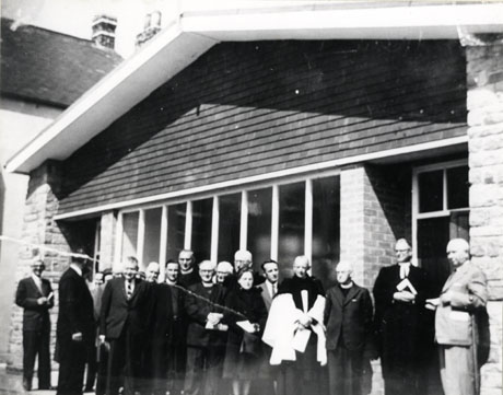 Photograph of nineteen people outside a building as in murt0099; the group of people include seven clergymen in dog collars; one of the clergy is in preaching bands and another is wearing a surplice; the photograph has been identified as Opening of New Methodist Church, Murton