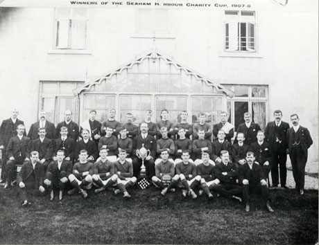 Photograph of seventeen men in football strip and nineteen other men, posed in front of a large building with a glass porch; in the middle of the group is a large trophy cup and a shield on which there are sixteen medals; a man on the front row is holding a ball with the following words on it : Murton Red Star 1907-08