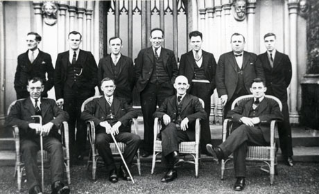 Photograph of eleven men posed before a nineteenth century Gothic wall and door, inside a building; seven of the men are standing behind the other four, who are sitting in basket chairs; two of the men sitting down are holding sticks; the men have been identified as Murton men at Conishead Priory, the Convalescent Home for Miners run by the Durham Miners' Association in Cumberland