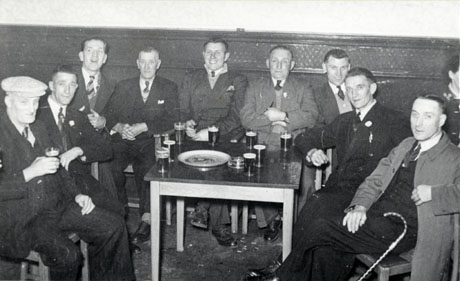 Photograph of nine men sitting round a table on which there are glasses of beer; the men are formally dressed in suits and ties; behind them is a wall on which there is a wainscot half way up; the photograph has been identified as recording Cup Final Celebrations, 1958