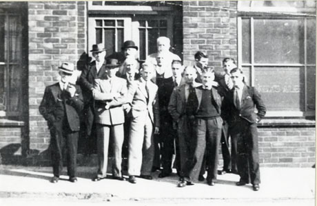 Photograph of a group of fourteen men standing outside the entrance of a public house; the word Ales can be seen engraved on a window of the building; the men are dressed formally in suits and ties; the photograph has been identified as showing a group of customers outside the Back Of The shaft public house
