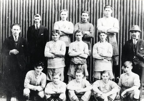 Photograph of a group of eleven young men and three men posed in front of a wall of corrugated iron; the young men are dressed in football strip and the other men in suits and ties; they have been identified as Footballers Outside Tin Chapel, Murton, 1920s