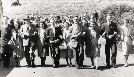 Photograph of five men and five women walking arm in arm along a road with a group of people and a hillside in the background; they are dressed in formal outdoor wear; the group has been identified as Murton Group on Visit to Northern Ireland, 1949