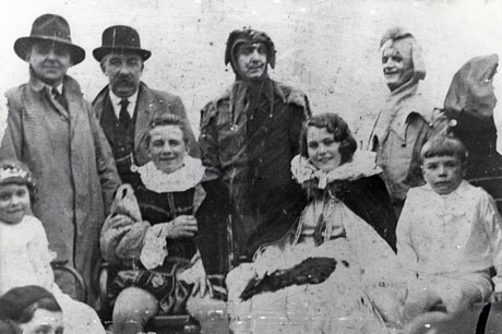 Photograph of a boy and girl aged approximately fourteen years, surrounded by two men dressed in hats and raincoats, two men dressed as jesters, a boy dressed as a page, and a small girl dressed as a maid of honour; the boy is dressed in Tudor costume of doublet and hose, and the girl is dressed in a long frock and a cloak; they have been described as Carnival King and Queen