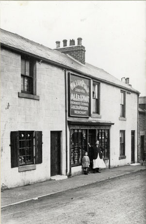 Photograph showing a terrace of three houses, the middle one of which is a shop; above the shop windows is a board reading: William Phillips Licensed To Retail Ale and Stout For Consumption Off The Premises Grocer And Provision Merchant; there are words above the windows which it is impossible to read; the contents of the windows cannot be seen; a man, wearing a dark jacket and a white apron, and a girl, aged approximately six years, are standing in front of the windows; the photograph has been identified as Phillipson Bailey Shop, Murton