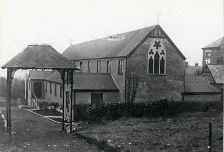 Photograph of the exterior of the East end of St. Joseph's Roman Catholic Church, Murton, showing the end of the building, a lower building on the south side of the building, a lych gate, a hedge immediately behind the church, and the roofs of adjacent buildings