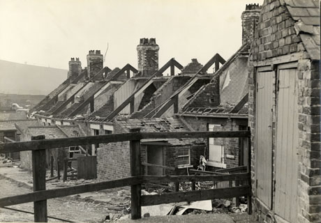 Photograph of houses, identified as South Crescent, Cold Hesledon, in the process of demolition; the roofs of the houses have been removed and the chimney stacks and roof timbers are still present; hills can be seen in the distance