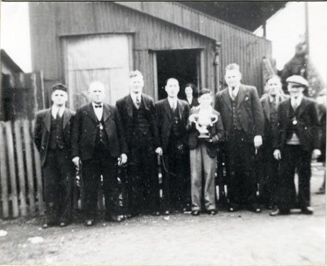 Photograph of a group of eight men standing outside a corrugated iron building, with a young boy in the middle of the group, holding a large trophy cup; a woman can be seen in the doorway of the building; the photograph has been identified as Presentation of Aged Miners' Cup, 1934