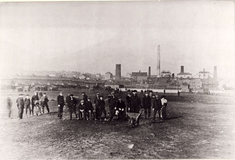 Photograph of a group of twenty four miners in the foreground, with more miners in the distance, in front of the buildings of Murton Colliery on the right of the picture; terraced houses can be seen on the left of the picture; one miner at the front of the picture is holding a wheelbarrow