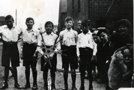 Photograph of five boys, aged approximately twelve years, wearing light shirts and dark shorts, standing in a row in the road, with a building on the right and a group of children, also on the right, watching the boys; the boy in the middle of the row, identified as H. Lowerson, is holding a star of five swords; the boys have been identified as Morton Juvenile Rapper-Sword Dancers