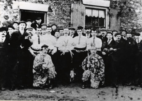 Photograph of five men in light shirts and dark trousers, surrounded by a crowd of approximately twenty five men; the middle man is holding a star of five swords; in front of the men are two men kneeling wearing patterned costumes and holding swords; the crowd is in front of a roughly built stone cottage, of which a window and a doorway can be seen ; the men have been identified as Murton Rapper-Sword Dance Team, circa 1904