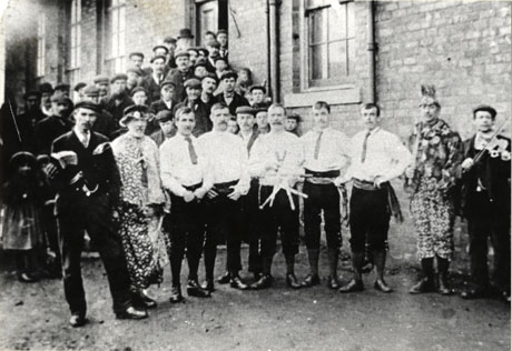 Photograph of five men in light shirts, knee breeches, and sashes posed in a row; a man in a highly patterned costume is standing at either end of the row of men; beside the men in patterned costume are men in ordinary suits holding an accordion and a violin; behind the men is a large brick building and a crowd of people standing on the steps leading up to its doorway; the men have been identified as Murton Rapper-Sword Dance team, circa 1904