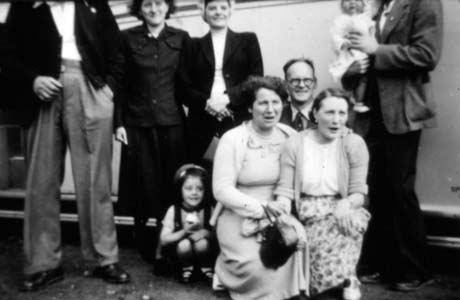 Photograph of two women, a man and a small girl, kneeling, with two men and two women standing behind them; the women appear to be singing; behind the group is the side of a bus; they have been identified as people from Murton on a bus trip
