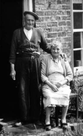 Photograph of an elderly man wearing trousers, a cardigan, and a cap, standing in the doorway of a house with his hand on the shoulder of an elderly woman, who is sitting on a chair wearing a pinafore and cardigan; they have been identified as Jimmy and Marjorie Lashley of Horden