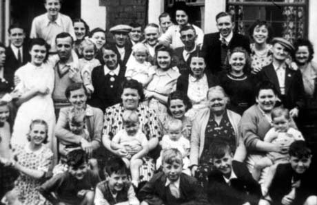 Photograph of a goup of approximately thirty men and women and thirteen children aged between one and ten years, posed in a group in front of the windows of a house; they have been identified as a group from Horden in Blackpool