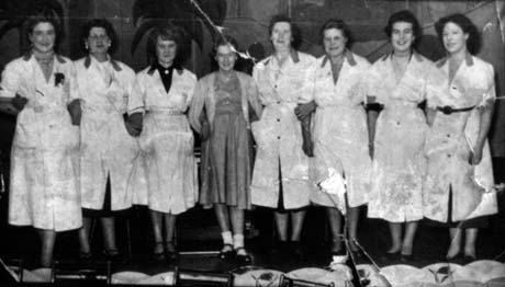 Photograph of seven women in uniform standing in a row; a woman wearing a dress and cardigan is in the middle of the row; they have been identified as Barmaids At Democratic Club, Murton