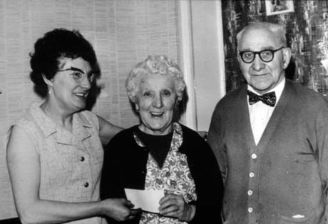 Photograph showing a middle-aged woman wearing a sleeveless dress with her arms round an elderly woman wearing a pinafore dress and cardigan; she is handing the woman a cheque; an elderly man wearing a cardigan and a bow tie is standing on the right; they have been identified as Jennie Raine and a couple whose house has burned down and who are the recipients of money raised at a charity show