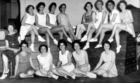 Photograph of eight women, wearing short skirts, sitting on a vaulting horse with their legs crossed; in front of them are five women sitting on the ground similarly dressed; a woman is sitting on a stool in front of a piano; they have been identified as members of a Keep-Fit Class in Horden