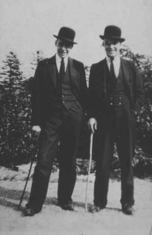 Photograph of two men dressed in suits and bowler hats and resting on canes; behind them are trees; the photograph has been identified as Conishead Home
