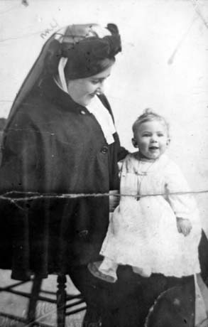 Photograph of a woman in a dark uniform of a cap and veil and cape and long skirt, sitting on a chair with an infant, wearing a frilly gown, sitting on her knee; she has been identified as Mrs. Garrett - 16-Year-Old Nurse Maid