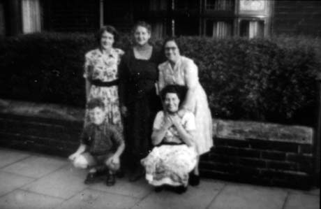 Photograph showing three women dressed in frocks standing in front of an indistinct background with a boy, aged approximately ten years, and a woman dressed in a frock squatting in front of them; they have been identified as Group at Knaresborough Road, Murton