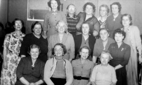 Photograph of seventeen women, wearing dresses and cardigans, posed in four rows in front of a wall in a building; they have been identified as British Legion Committee, Murton